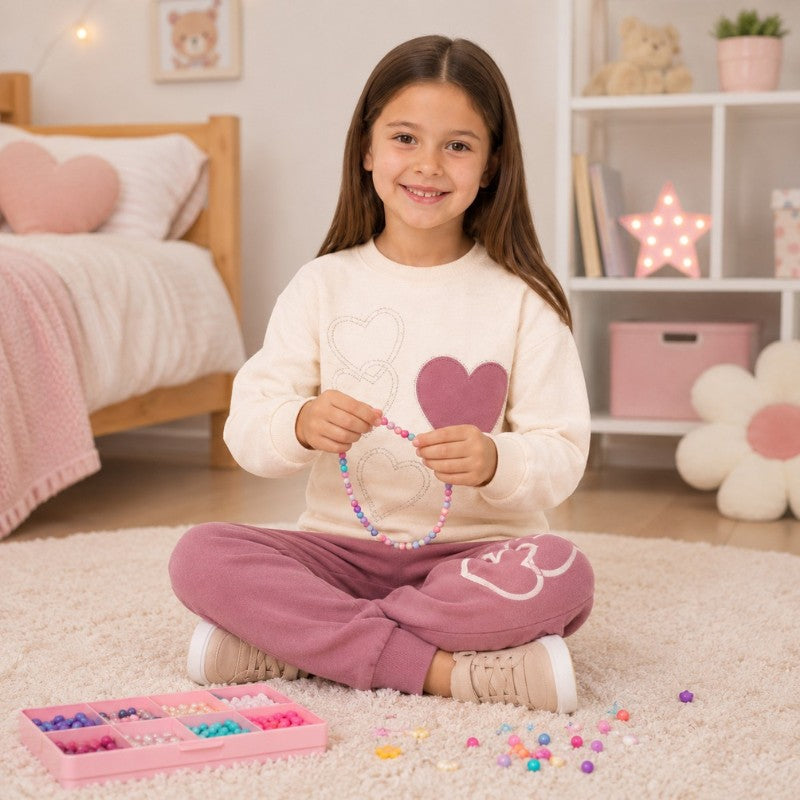 Young girl wearing a cream heart-print sweatshirt and dusty pink jogger pants while making a colorful bead necklace.