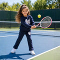 Young girl wearing a navy floral tracksuit and white sneakers while playing tennis on an outdoor court.