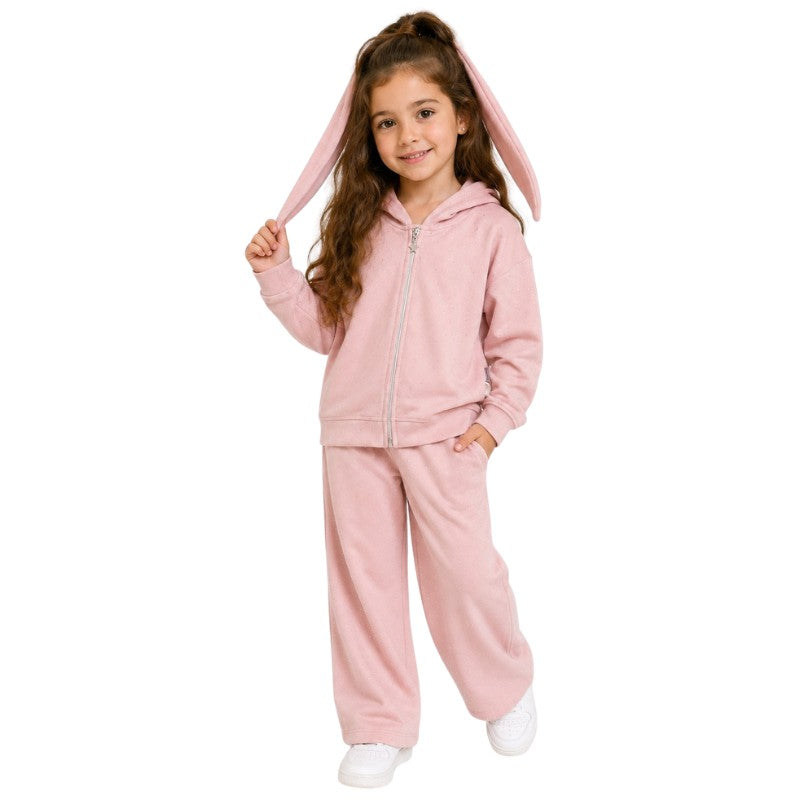 Young girl wearing a pink bunny-ear hoodie tracksuit set in a bright cosy room.