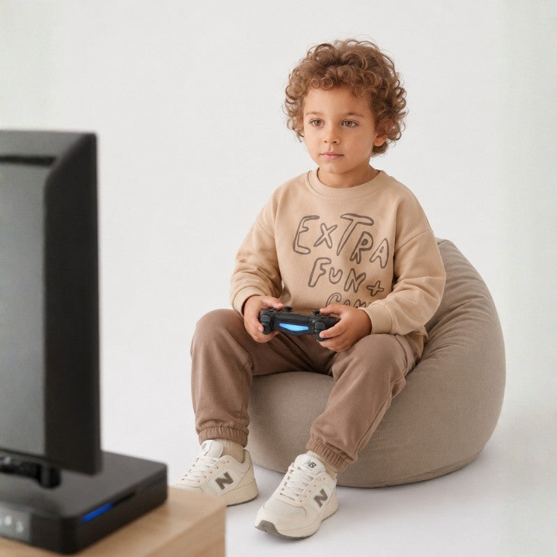 Young boy wearing a beige graphic sweatshirt, brown jogger pants, and white sneakers while playing video games.