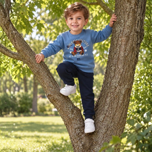Young boy wearing a light blue teddy bear sweatshirt and navy pants climbing a tree outdoors