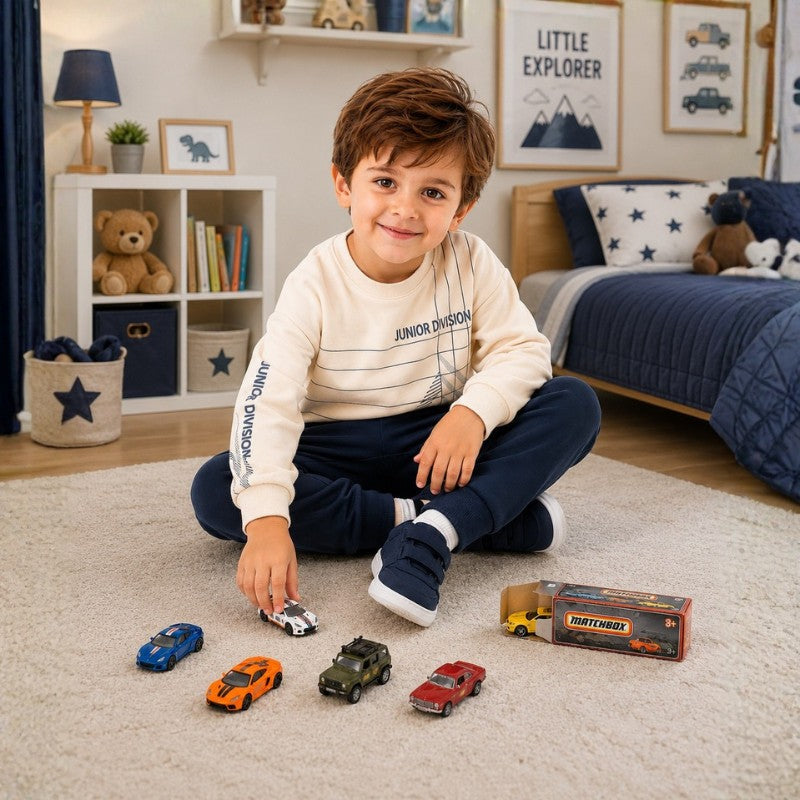 Young boy wearing a cream graphic sweatshirt and navy jogger pants while playing with toy cars in his bedroom.